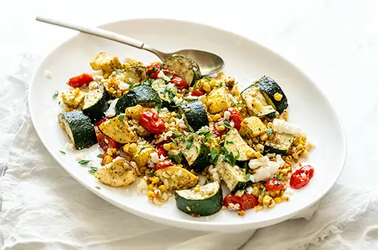 Platter of roasted summer vegetables with zucchini, yellow squash, corn, cherry tomatoes, parsley, and feta cheese, served with a spoon on a white linen backdrop.