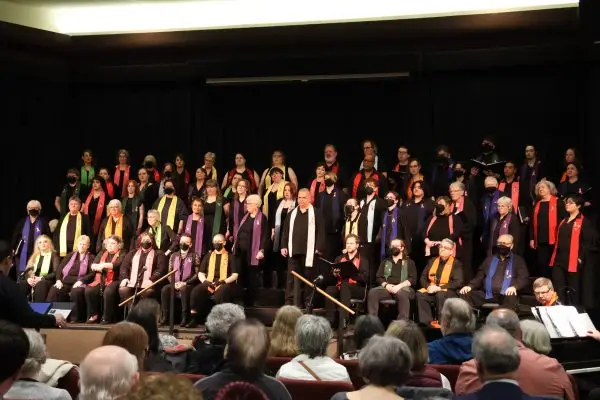 Members of Confluence: Willamette Valley LGBT Chorus perform on stage before a seated audience. Singers of varying ages stand on risers wearing black attire with colorful rainbow scarves, celebrating community, diversity, and LGBTQ+ pride through music.